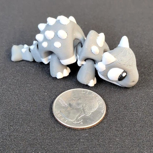 Gray articulated 3D printed Ankylosaurus with white armor and tail club beside a quarter for scale