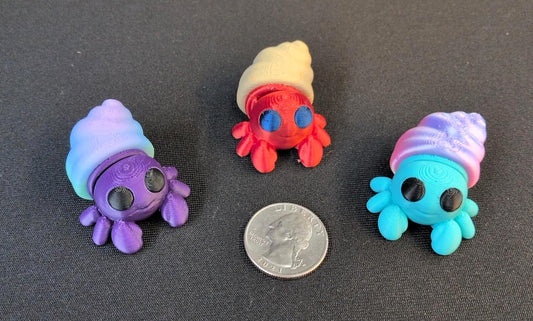Three colorful 3D printed hermit crabs with shells and a U.S. quarter for scale